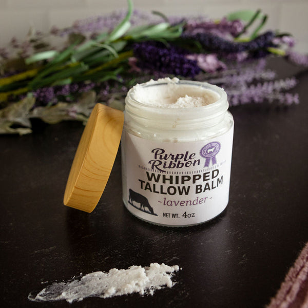Jar of Purple Ribbon Whipped Tallow Balm on a dark surface with lavender flowers in the background