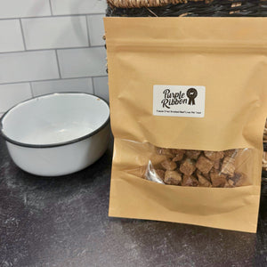Kraft paper bag with pet treats labeled 'Purple Ribbon' on a dark surface with a wicker basket and white bowl in the background.