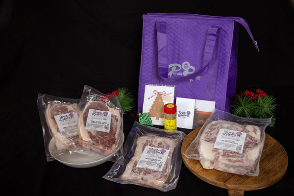 A packaged meat gift set displayed on a black background, including three vacuum-sealed cuts of beef with labels, a purple reusable bag with a white logo, small gift cards, a yellow bottle, and greenery with red berries.