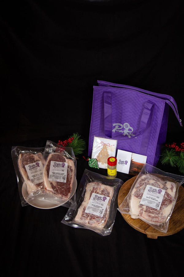 A packaged meat gift set displayed on a black background, including three vacuum-sealed cuts of beef, a purple reusable tote bag, small seasoning bottles, and a few packaged accessories. The labels show the brand Beef Heritage.
