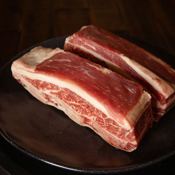 Two pieces of raw beef short ribs on a dark surface