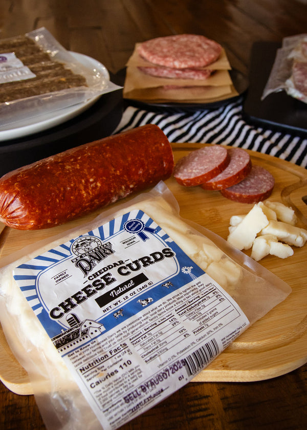 Packaged cheese curds with a salami and sliced meat on a wooden board.