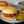 Cargar imagen en el visor de la galería, A plated sloppy joe sandwich with a sesame bun, served with fries and pickles on a dark tabletop, with additional plates and a bowl blurred in the background.
