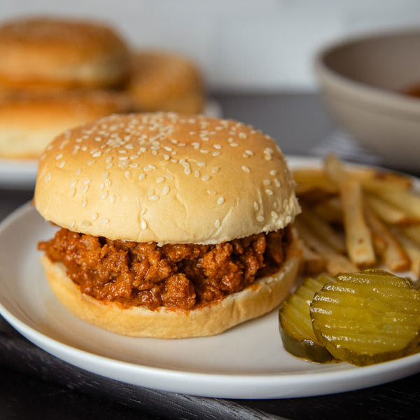 A plated sloppy joe sandwich with a sesame bun, served with fries and pickles on a dark tabletop, with additional plates and a bowl blurred in the background.