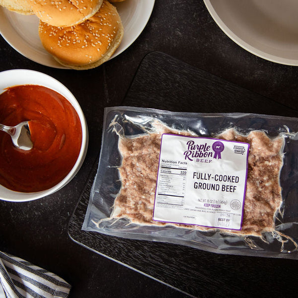 A vacuum-sealed package of Purple Ribbon Fully Cooked Ground Beef placed on a dark surface, surrounded by a bowl of red sauce, burger buns, and a striped cloth.
