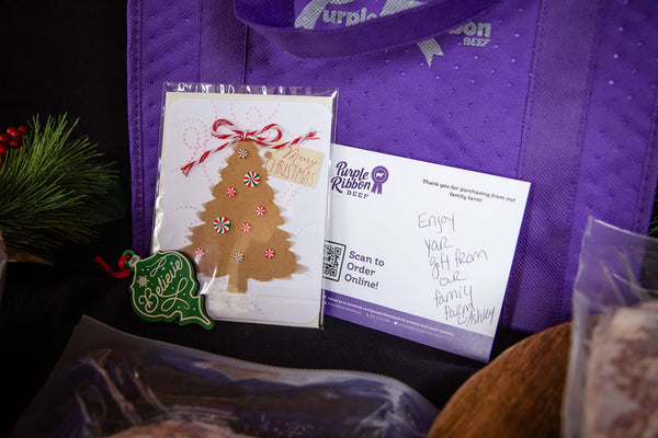 A purple reusable shopping bag with white text, alongside a Christmas tree ornament card, a white order card from Purple Ribbon, and a green holiday ornament, arranged on a dark surface.