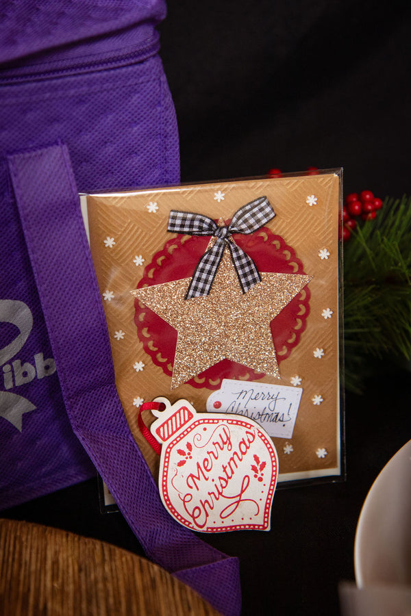 A packaged Christmas greeting card or gift wrap featuring a gold star, red circular pattern, white snowflakes, a black-and-white gingham bow, and a hanging red-and-white ornament tag that reads 'Merry Christmas.' It is photographed beside a purple tote bag with partial branding visible, with dark background and small greenery/red berry props around it.